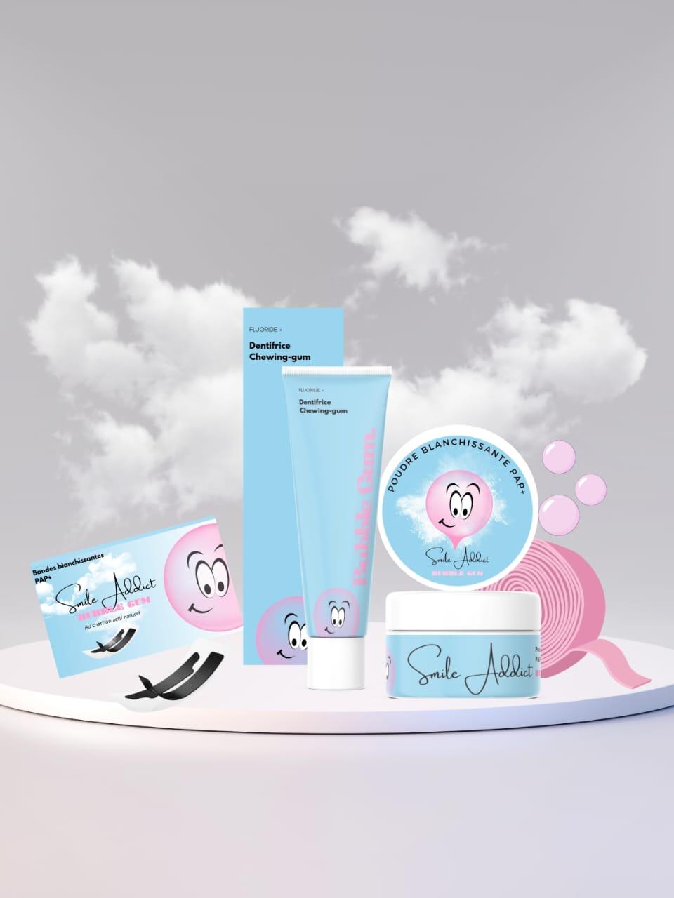 A display of Smile Addict's Pack Xtreme Blancheur Bubble Gum—Bandes PAP+, charbon, poudre, et dentifrice—features blue and pink packaging with a smiling cartoon face, set against a cloudy background.